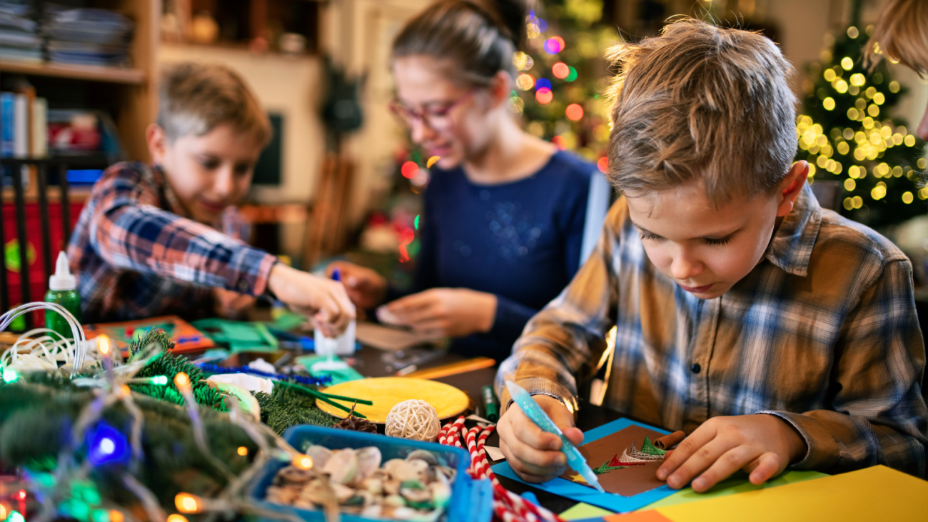 Children creating holiday crafts at a festive table, surrounded by colorful supplies, twinkling lights, and holiday decorations, with a Christmas tree glowing softly in the background.