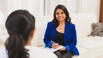 Yolanda, a parenting and relationship expert, smiling warmly during a one-on-one coaching session, seated on a white couch with a notepad in hand, creating a safe and supportive space for parents.