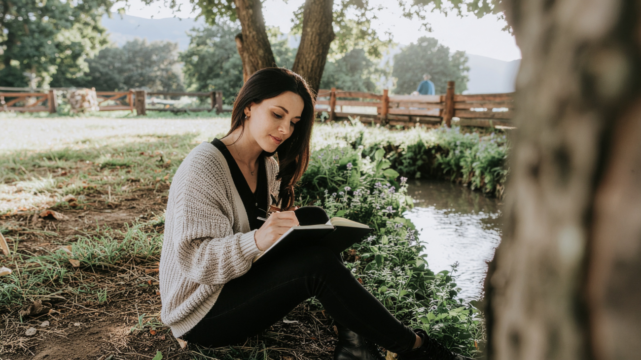 young woman writing on her notebook surrounded by a beautiful view of trees, water, and bridge