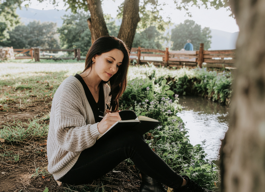 young woman writing on her notebook surrounded by a beautiful view of trees, water, and bridge