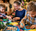 Children creating holiday crafts at a festive table, surrounded by colorful supplies, twinkling lights, and holiday decorations, with a Christmas tree glowing softly in the background.