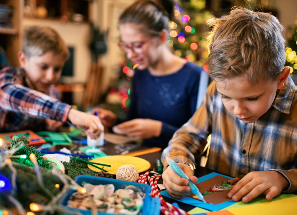 Children creating holiday crafts at a festive table, surrounded by colorful supplies, twinkling lights, and holiday decorations, with a Christmas tree glowing softly in the background.